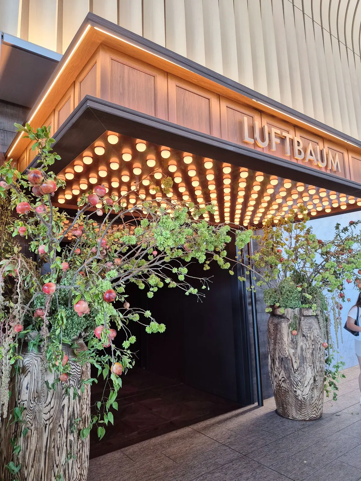 木製の柱に実った赤い果実の装飾が並ぶ入り口。上部には「LUFTBAUM」のネオンサインと、多数の電球が放射状に配置された装飾がある。