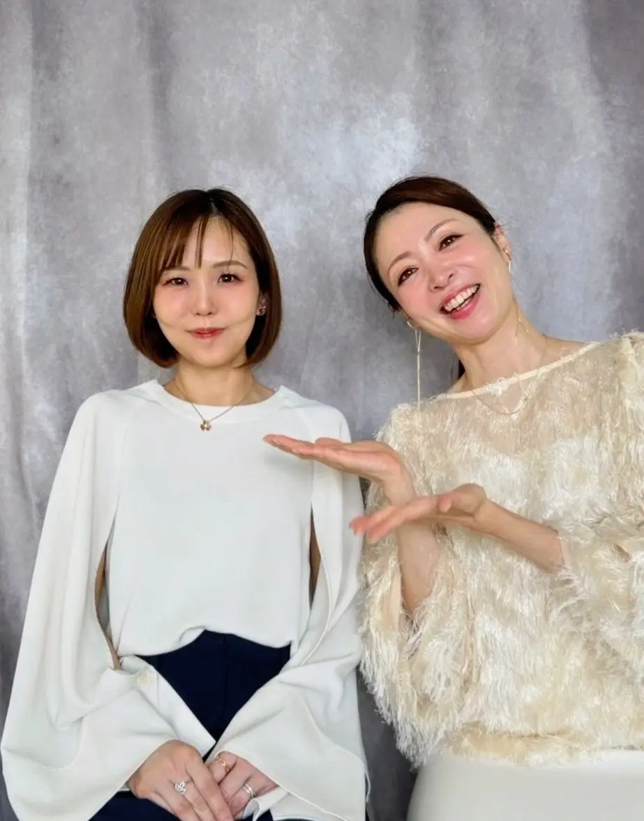 二人の女性がグレーの背景に並んで座っている。左の女性はショートカットで白い上着を着て、右の女性はロングヘアでふわふわのベージュの服を着て、笑顔で手を出している。