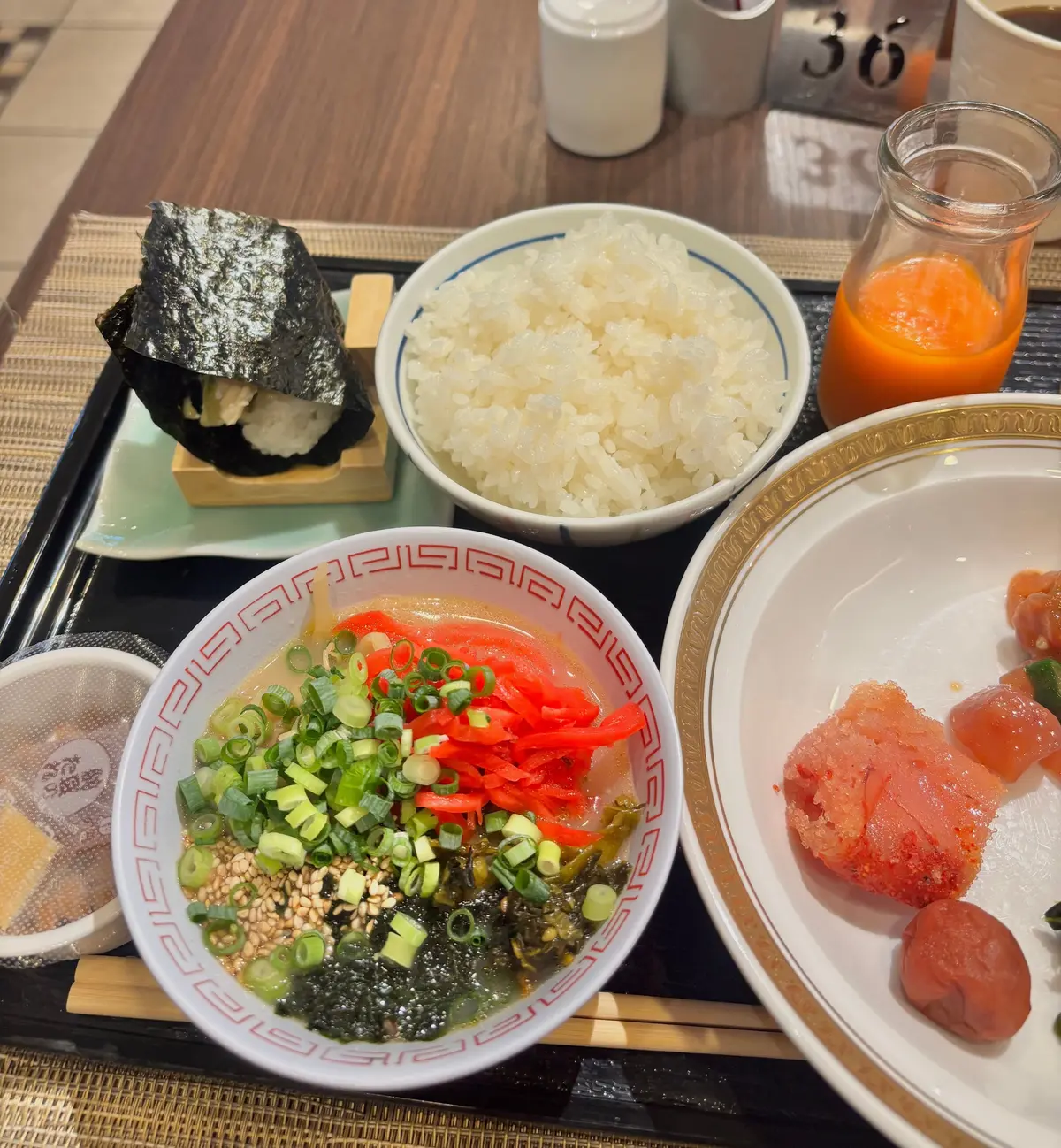 黒いお弁当箱に盛り付けられた和食。ご飯、おにぎり、海苔の佃煮、だし巻き卵、そしてカットフルーツ。右には飲み物が置かれている。