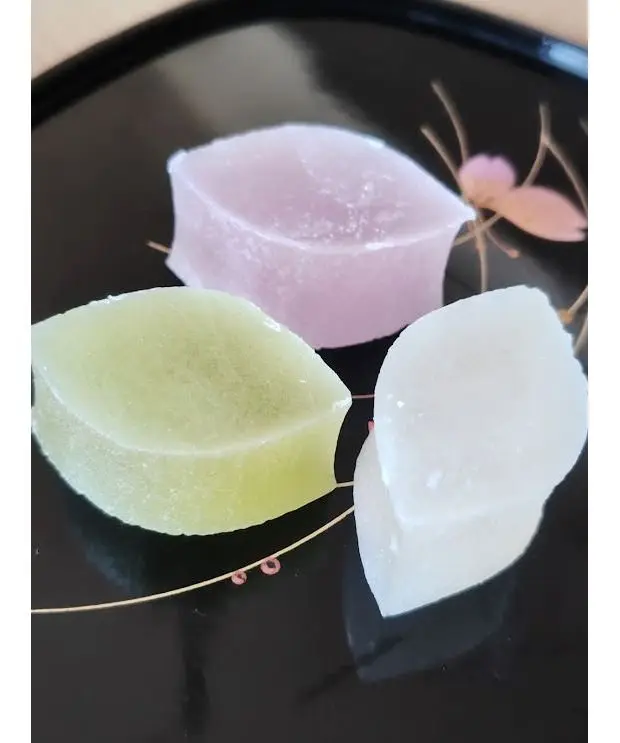 黒いお皿に、ピンク、緑、白の3つの和菓子が並んでいる。それぞれが四角くカットされ、異なる色合いを見せている。お皿には金色の模様と、桜の花の絵柄が描かれている。