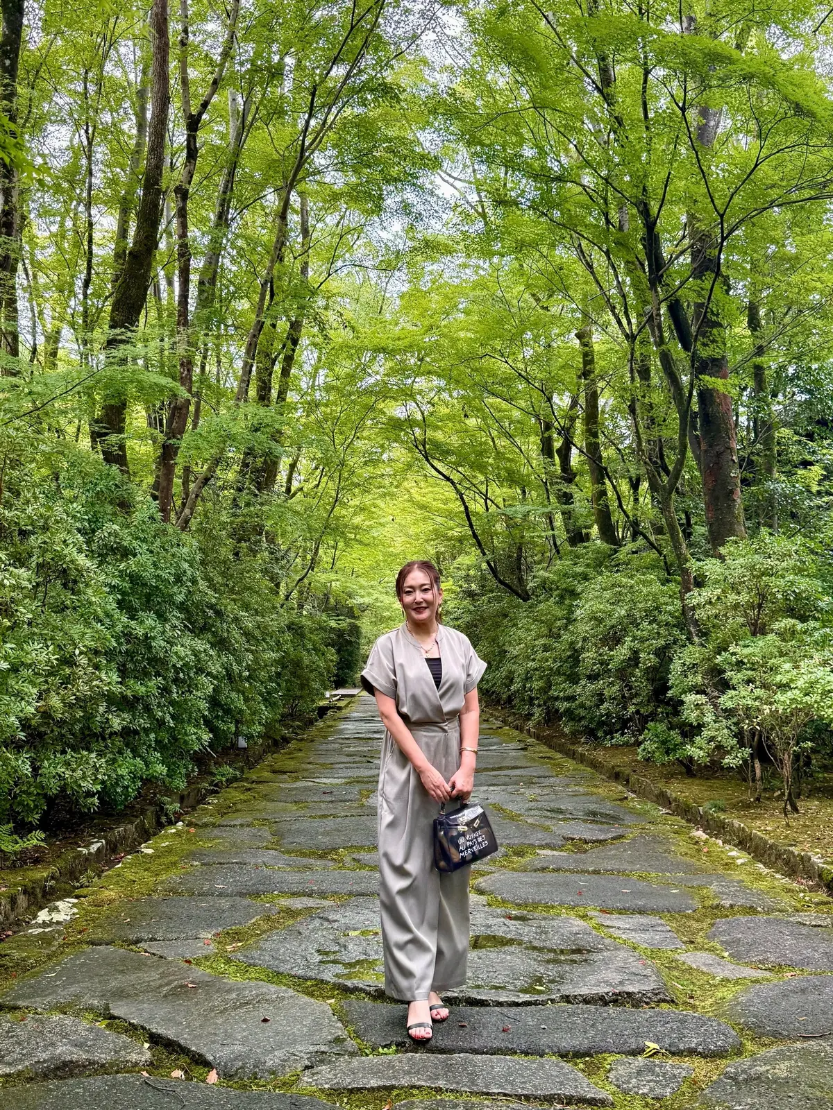 石畳の道の中央に立つ女性。グレーのオールインワンの服を着ており、右手に黒いゼリーバッグを持っている。背景には緑豊かな木々が茂り、自然のトンネルのようになっている。