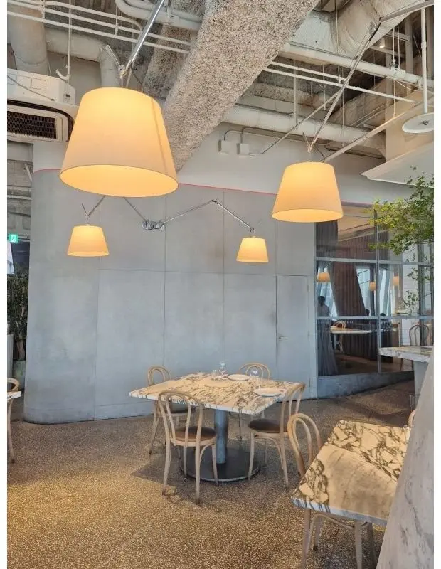 モダンなカフェの内装で、円形のテーブルと木製の椅子が配置され、複数のペンダントライトが天井から下がっている。壁はコンクリート調で、右側にはガラスの仕切りがあり、植物が置かれている。