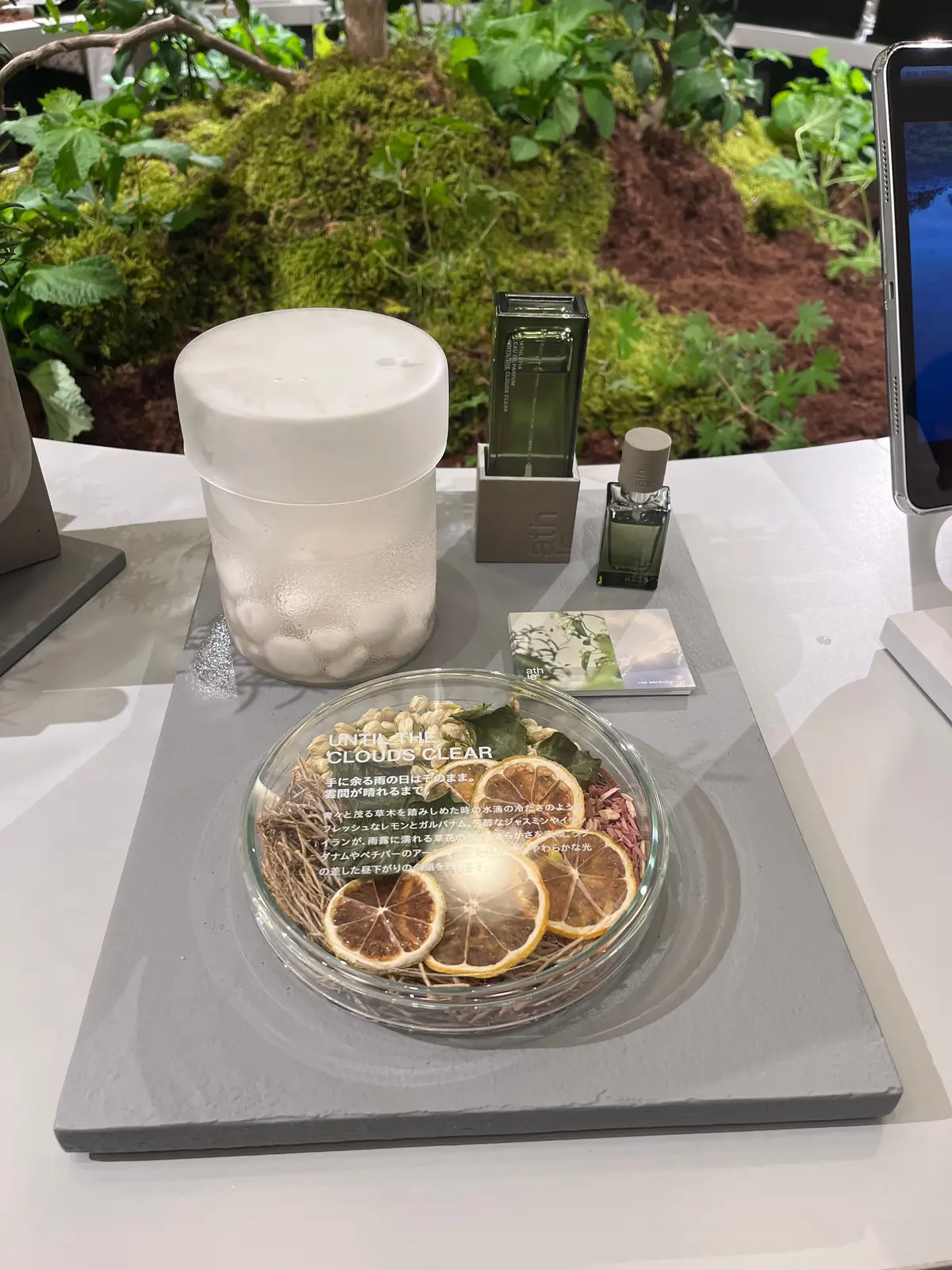 白いディフューザー、緑色のボトル、オレンジのスライスが置かれた展示台。背景には植物が茂り、右端には黒いタブレットが映っている。台には「UNIZUE CLOUDS CLEAR」と書かれたカードが置かれている。