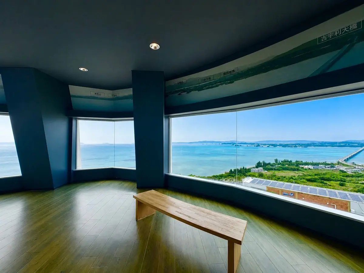 青い壁の部屋で、大きな窓から海と陸地が広がる景色が見える。床は木製で、窓の前に木製のベンチがある。天井には丸い照明が取り付けられている。