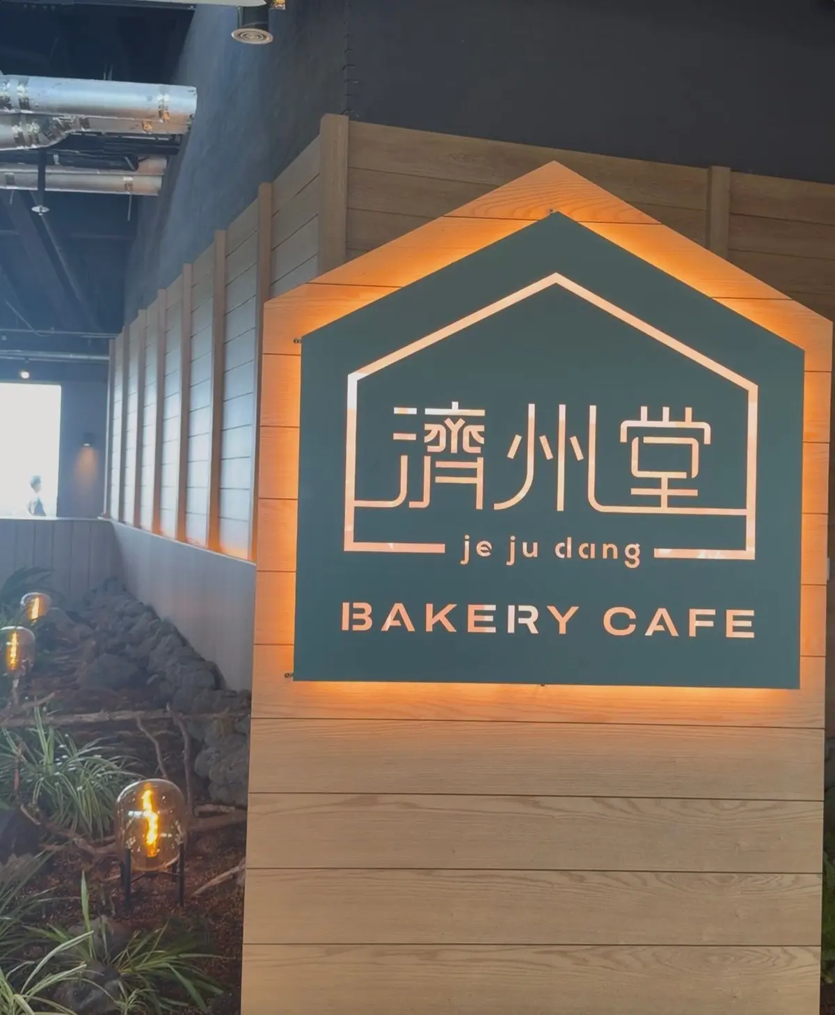木製の柱に設置された、緑色の屋根付きの看板。中央には漢字で「清州堂」、その下に「je ju dang」、さらに「BAKERY CAFE」と書かれている。看板の周囲は明るくライトアップされている。