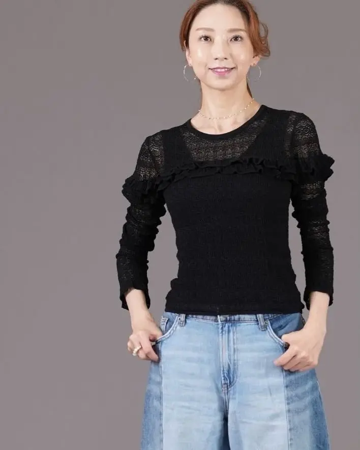 黒いニットトップスにブルーのワイドデニムを着用した女性が立っている全身写真。茶色の髪を後ろでまとめ、控えめな微笑みを浮かべている。
