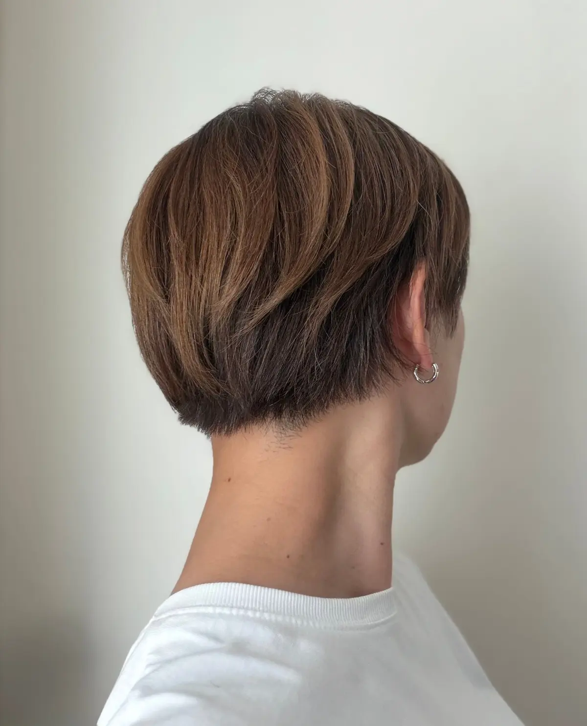 茶色のショートヘアの女性。後頭部から首筋にかけて見え、耳にはシルバーのフープピアスをしている。白いTシャツを着用。