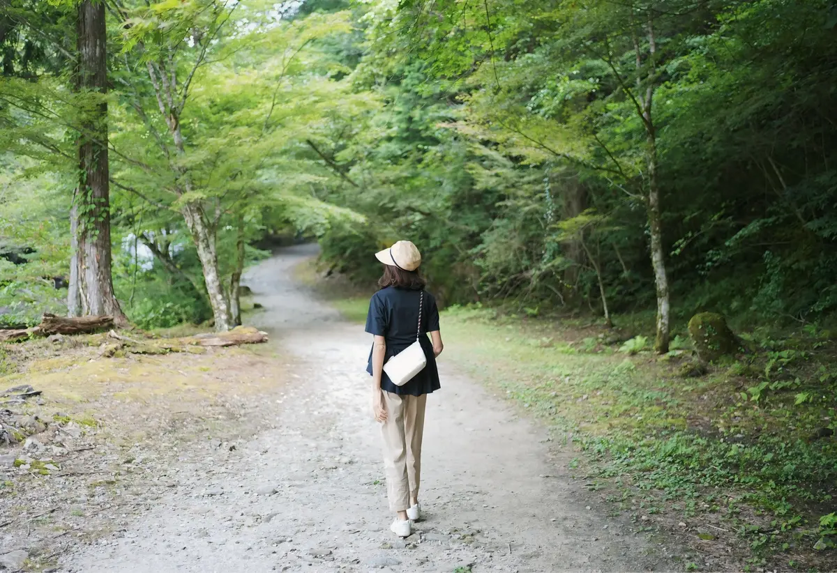 緑豊かな木々の間の小道に立つ女性。麦わら帽子をかぶり、紺色のトップスにベージュのパンツ、白いスニーカーを着用。白いショルダーバッグを斜め掛けし、前方を向いて歩いている。