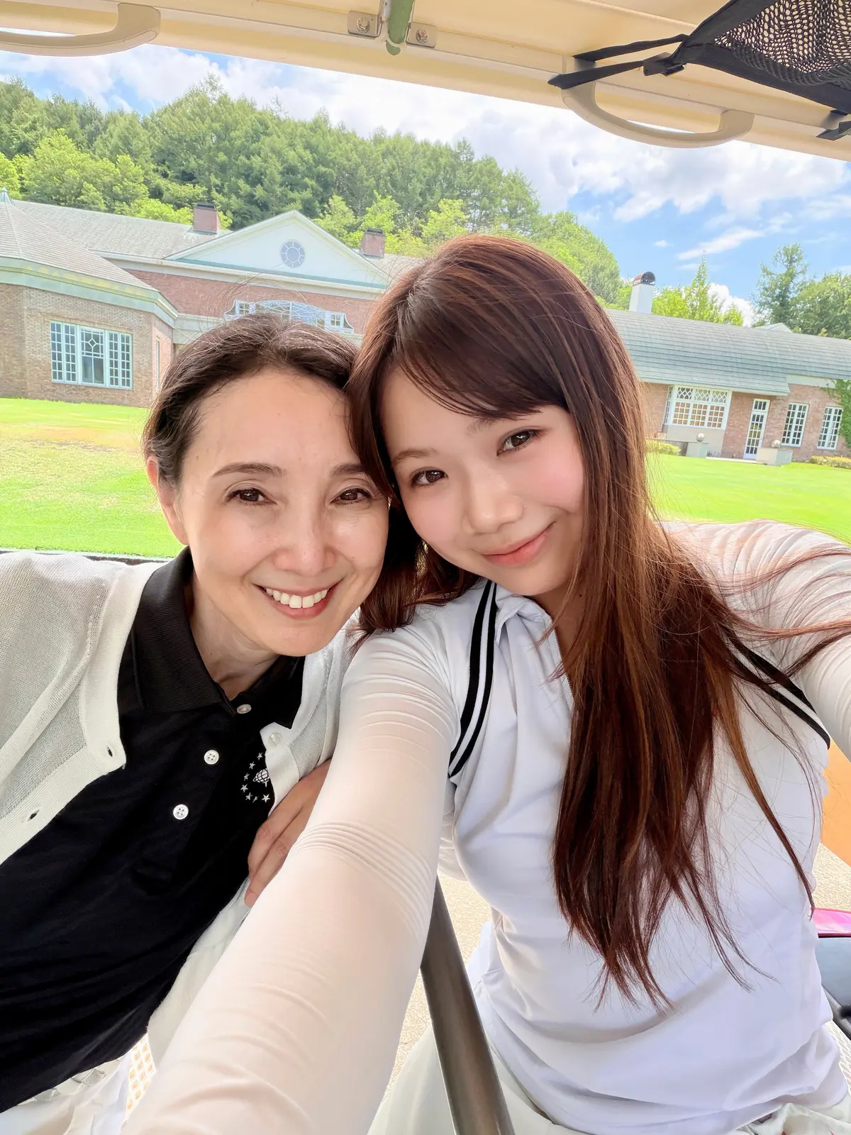 ゴルフカートに乗った2人の女性が笑顔で自撮りしている写真。背景には芝生と建物が見える、晴れた日の屋外。左の女性は黒と白の服にグレーのジャケット、右の女性は白いシャツを着ている。