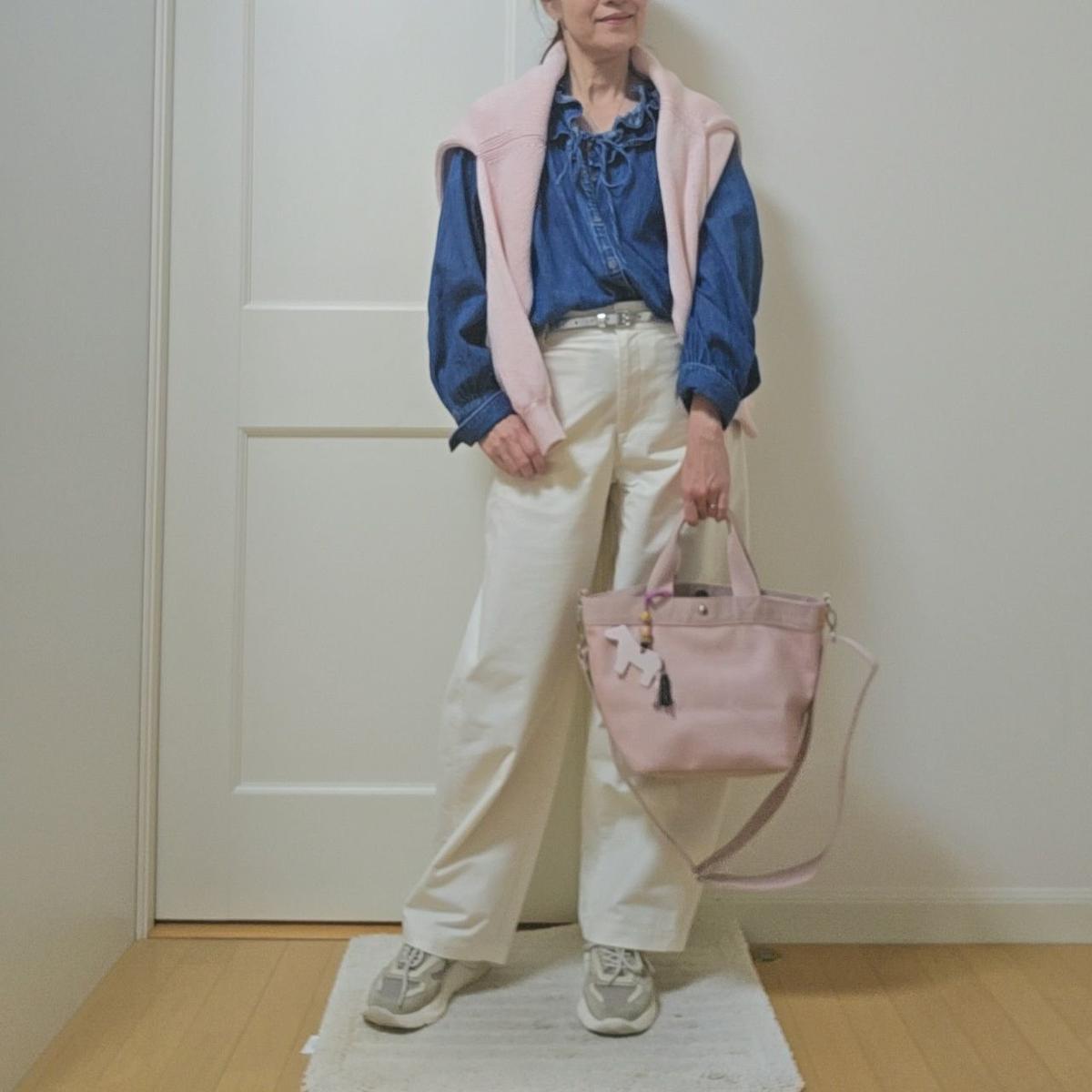白いパンツ、デニムシャツ、ピンクのカーディガンを着た女性。ピンクのバッグを持ち、白いスニーカーを履いている。背景は白いドア。