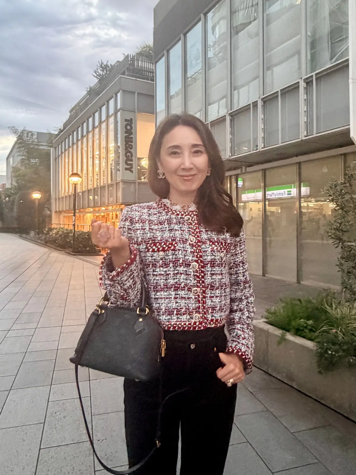 長めの茶髪の女性が、黒いバッグを肩にかけ、笑顔でポーズをとっている。背景には、TONI&GUYの看板がある建物が見える。