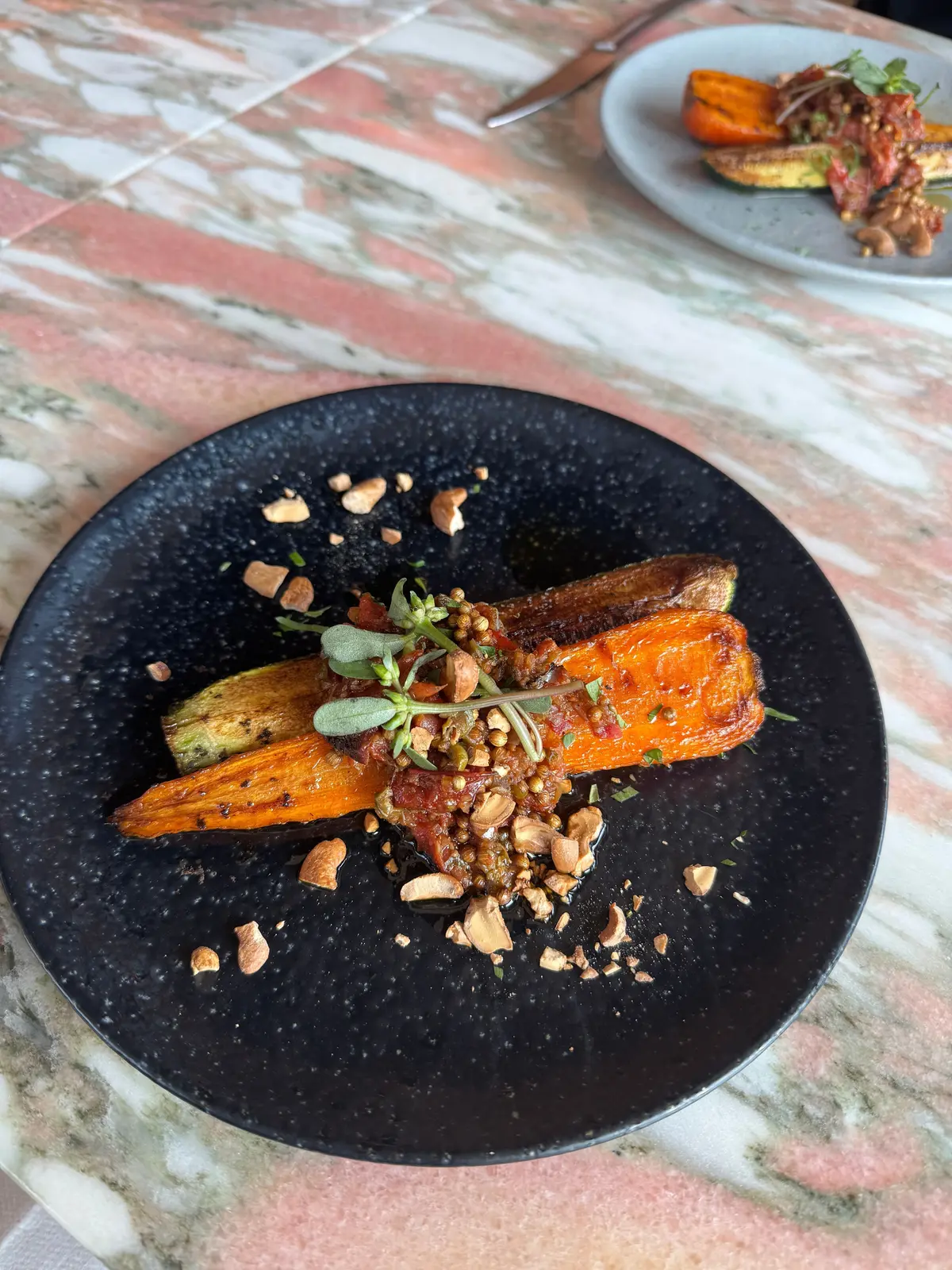 黒い皿に盛り付けられた、ローストしたカラフルな野菜。オレンジ、紫、緑色の人参が並び、ソースと砕いたナッツ、ハーブが添えられている。背景にはマーブル模様のテーブルと、別の皿が見える。