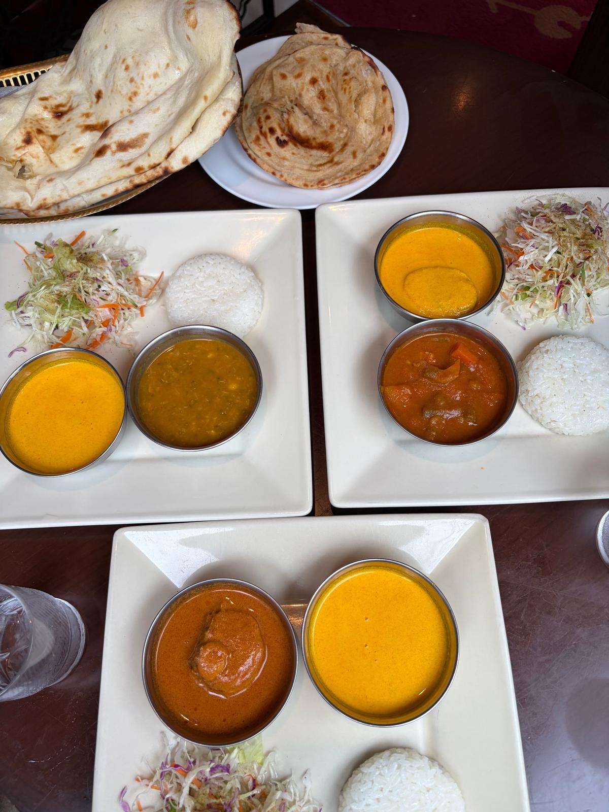 木製のテーブルに置かれた、インド料理の盛り合わせ。左上にはナンが2枚、右には白米とコールスロー、様々なカレーが小鉢に盛られている。