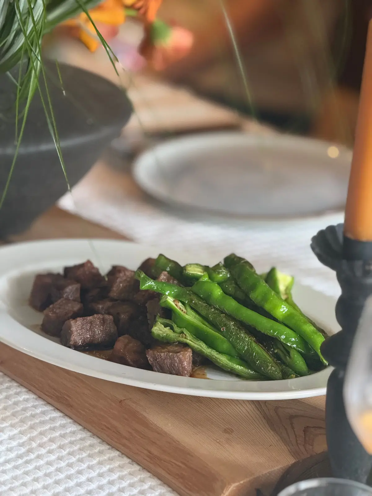 白磁の楕円皿に盛り付けられた、牛肉と緑色のピーマンの炒め物。牛肉は角切りで、ピーマンは縦にカットされている。木製のトレイに皿が置かれ、背景にはキャンドルと花が見える。