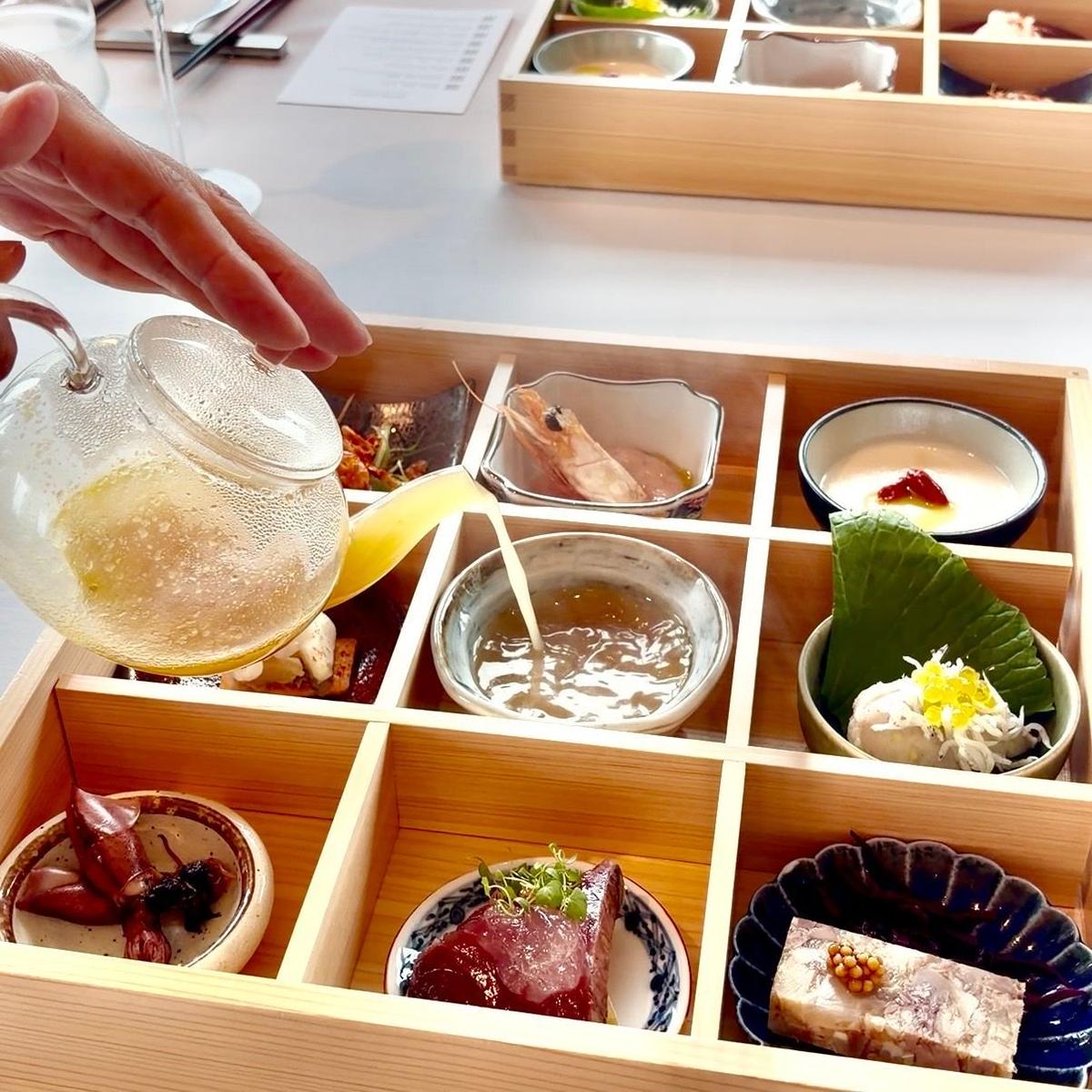 松花堂スタイル。木製のトレイに並べられた９種類の豆皿に多様な料理が盛り付けられている。一つの豆皿にだし茶を注いでいる。