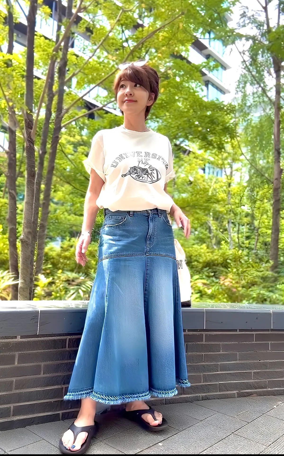 ベージュの大学ロゴTシャツとデニスカートを着た女性が、緑豊かな木々のある屋外で、壁に寄りかかって立っている。
