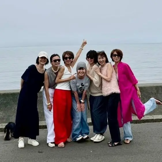 7人の女性と1人の男性が海辺の壁に寄り添って並んでいる。女性たちはさまざまな服装で、帽子やサングラスを着用。男性はミッキーマウスのTシャツを着ている。海が背景に見える。7人で集合写真。