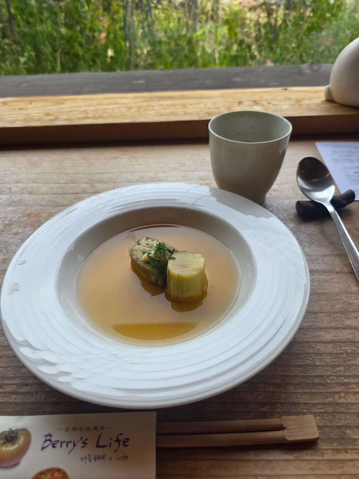白い皿に盛り付けられた、透明なスープと、ハーブで包まれた緑色の食材。右には茶色のマグカップとスプーン、手前には「Berry's Life」と書かれたメニューが置かれている。木製のテーブルと窓からの自然光が背景。