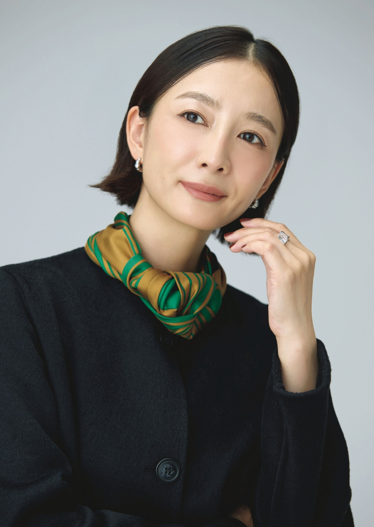 黒いジャケットに、緑と黄色の模様のシルクのスカーフを巻いた女性。短くカットされた黒髪で、控えめな微笑みを浮かべている。右手を頬にあて、指には大きな指輪が光っている。背景は白。