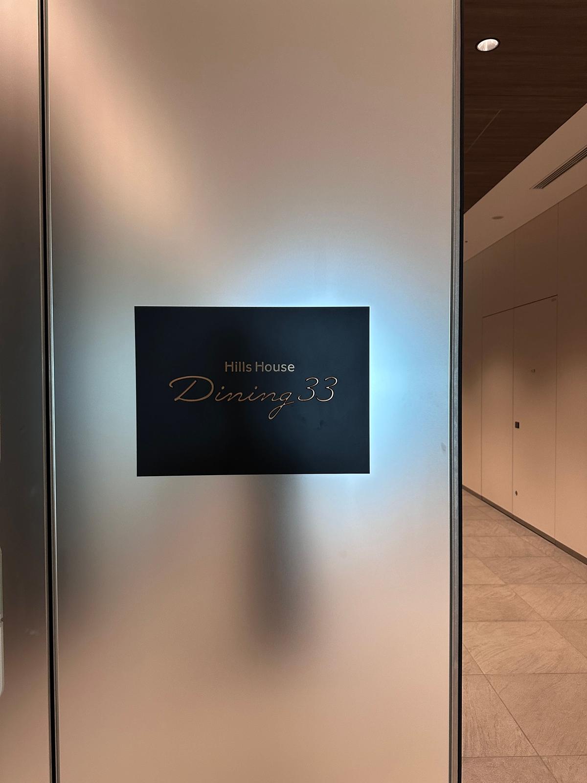Hills House Dining 33 と表示された黒い正方形のサインが、光沢のある壁に設置されている。右側には、ベージュ色の壁と石張りの床が見える。