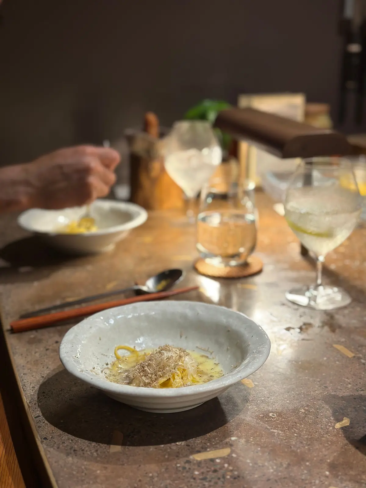 陶器のボウルに入ったパスタと、グラスに入った飲み物。テーブルには箸とスプーンが置かれ、背景には人物の手や他の食器が見える。