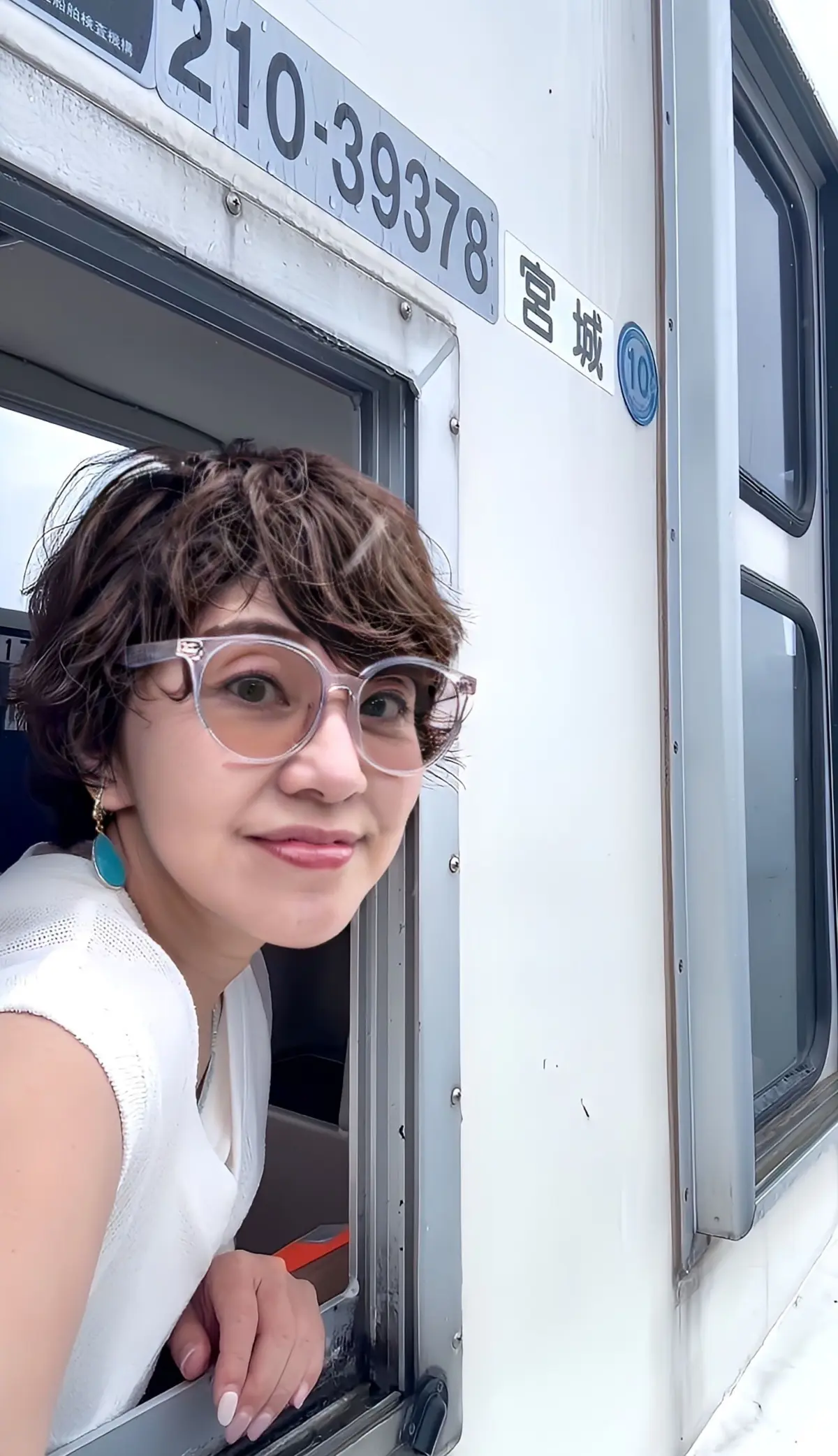 白いシャツを着た女性が、電車の中からこちらを見ている。茶色の短髪で、縁なしの眼鏡をかけている。背景には、電車の側面と番号が書かれたプレートが見える。