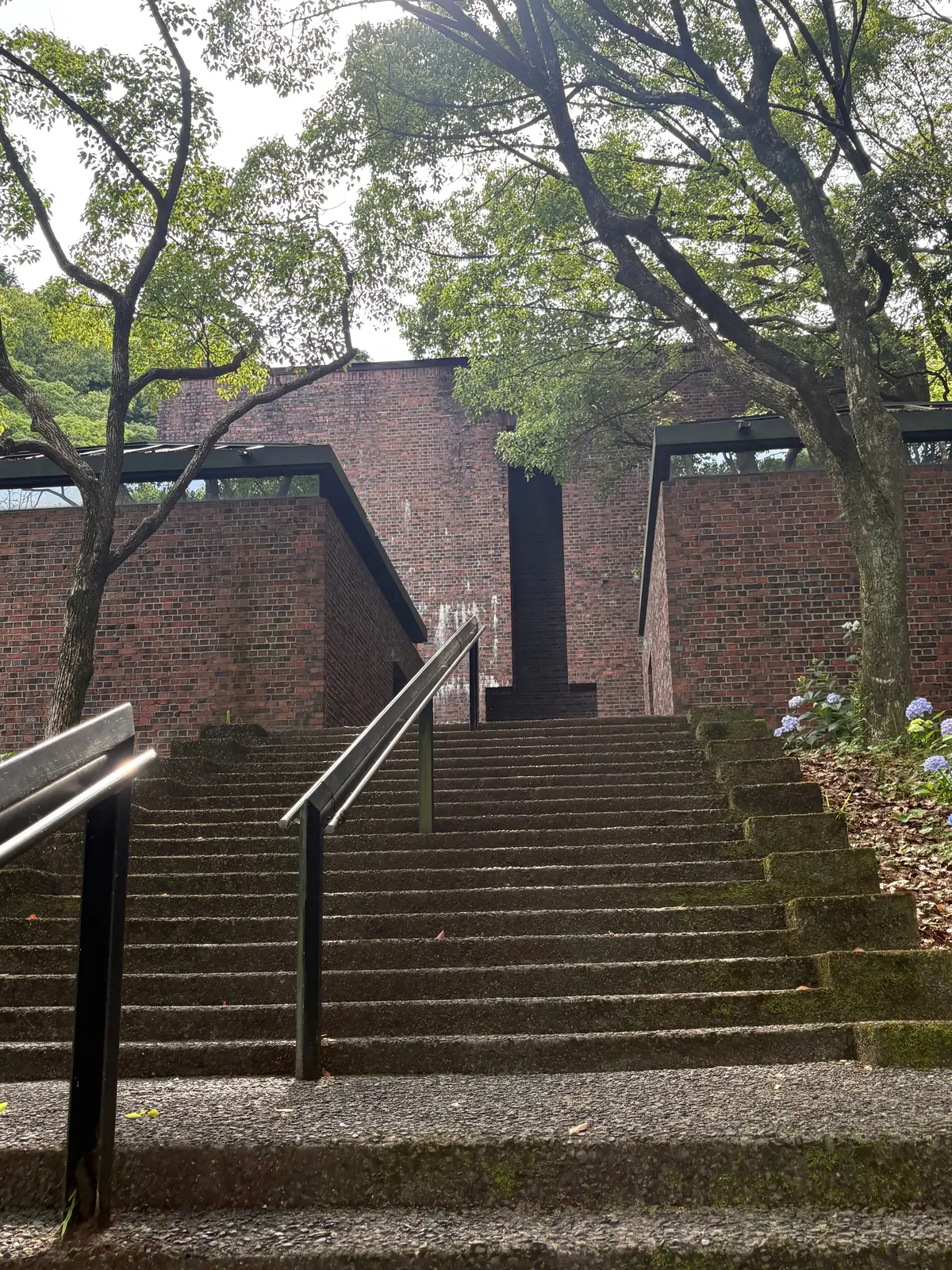 レンガの建物に続く石段と手すり、周囲に木々や青い花が咲く緑豊かな環境