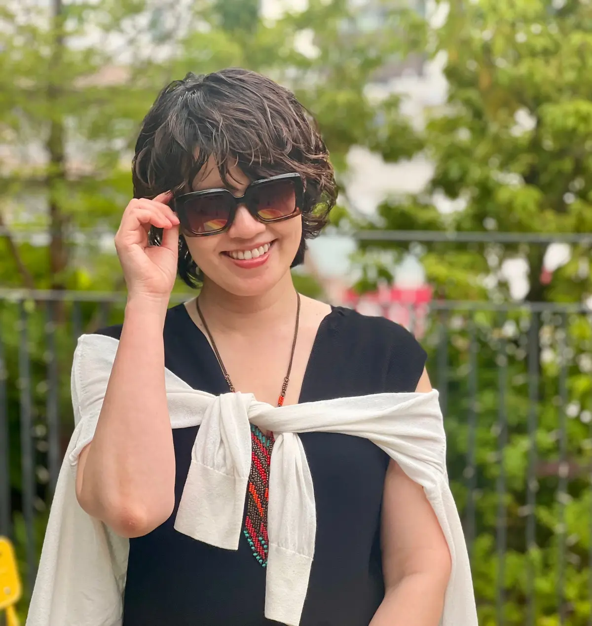 女性が黒いサングラスをかけ、白いリボンが付いた黒いドレスを着て、屋外で微笑んでいる。背景には緑の木々が見える。
