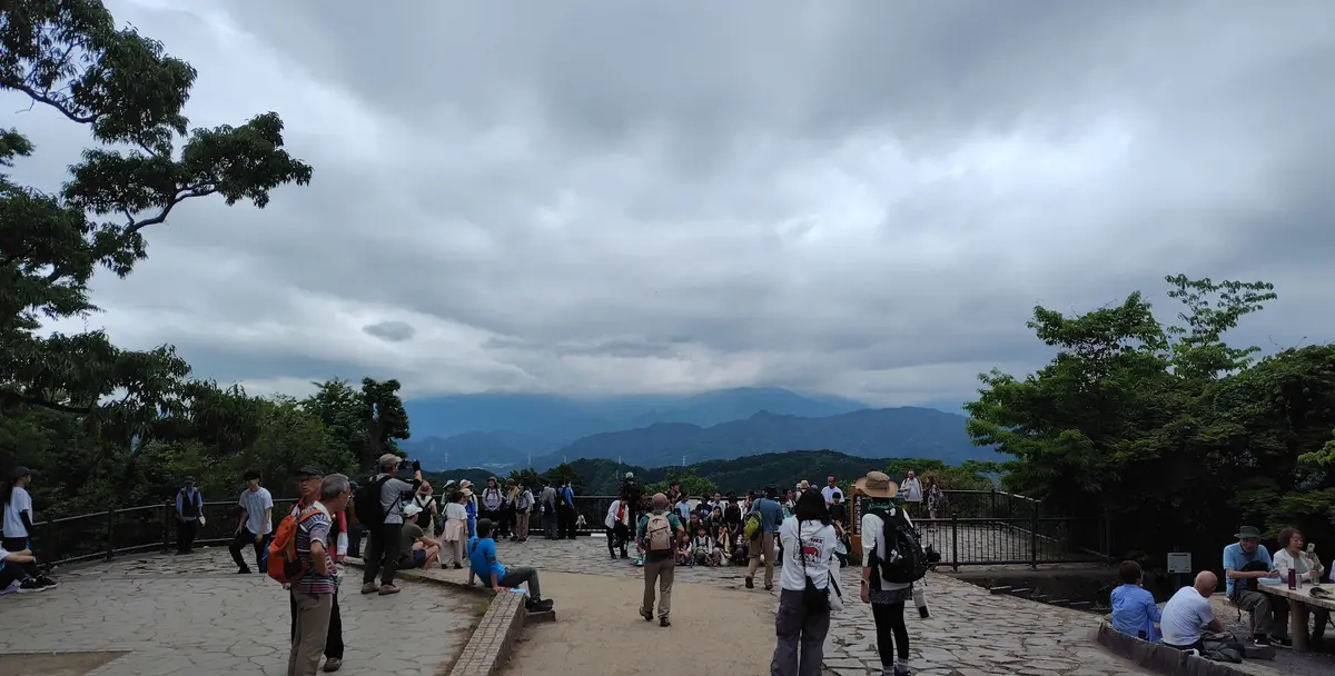 高尾山頂の展望台で多くの観光客が集まり、石畳の通路を歩きながら山並みを見上げています。