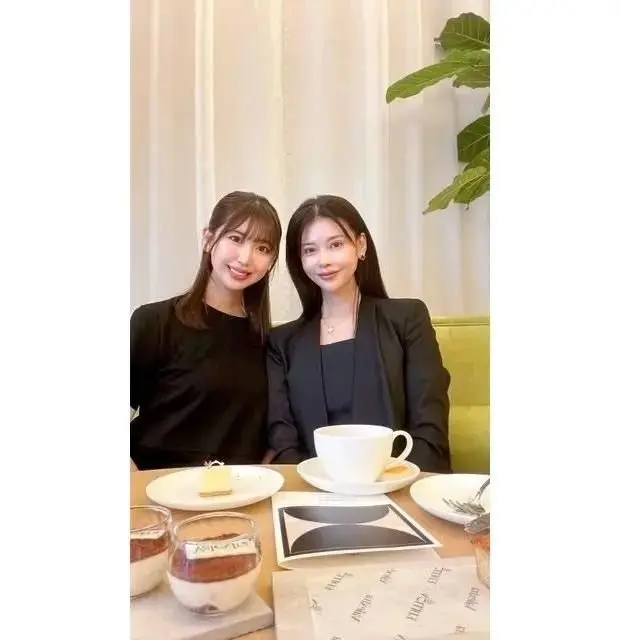 カフェでデザートを楽しむ2人の女性。左は茶色のショートヘア、右は黒髪でスーツを着ている。テーブルにはカップやデザートが置かれ、背景には白いカーテンと観葉植物が見える。