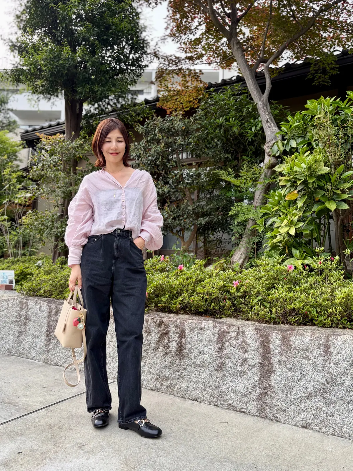 ピンクの長袖シャツと黒いデニムパンツを着た女性が、緑豊かな庭園で立ち、笑顔でカメラを見つめている。左手にベージュのトートバッグを持ち、黒いローファーを履いている。