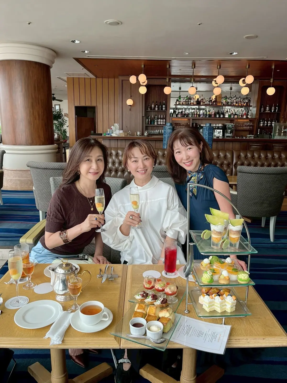 3人の女性がテーブルに座り、シャンパンを持って笑っている。テーブルにはティーカップ、グラス、フルーツやデザートの盛り合わせが置かれている。背景にはバーカウンターと装飾的な照明がある。