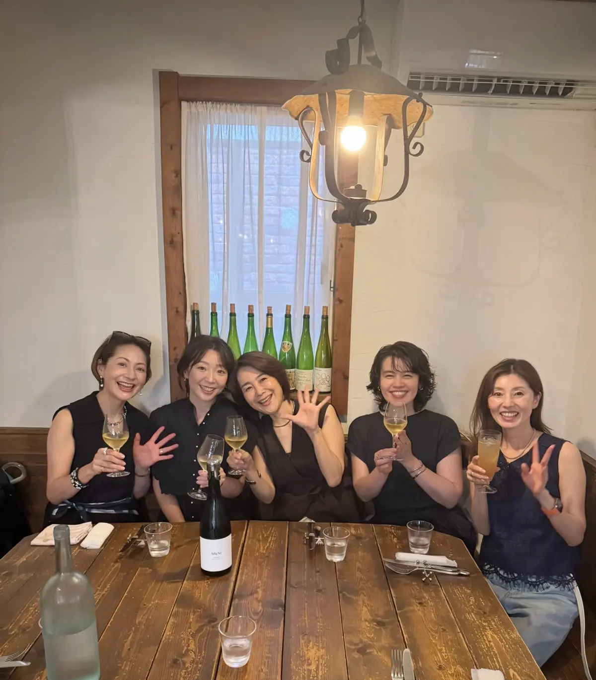 木製のテーブルを囲み、5人の女性がグラスを片手に笑顔で写っている。背景には窓と装飾的な照明器具が見える。テーブルにはワインボトルやグラス、食器類が並んでいる。