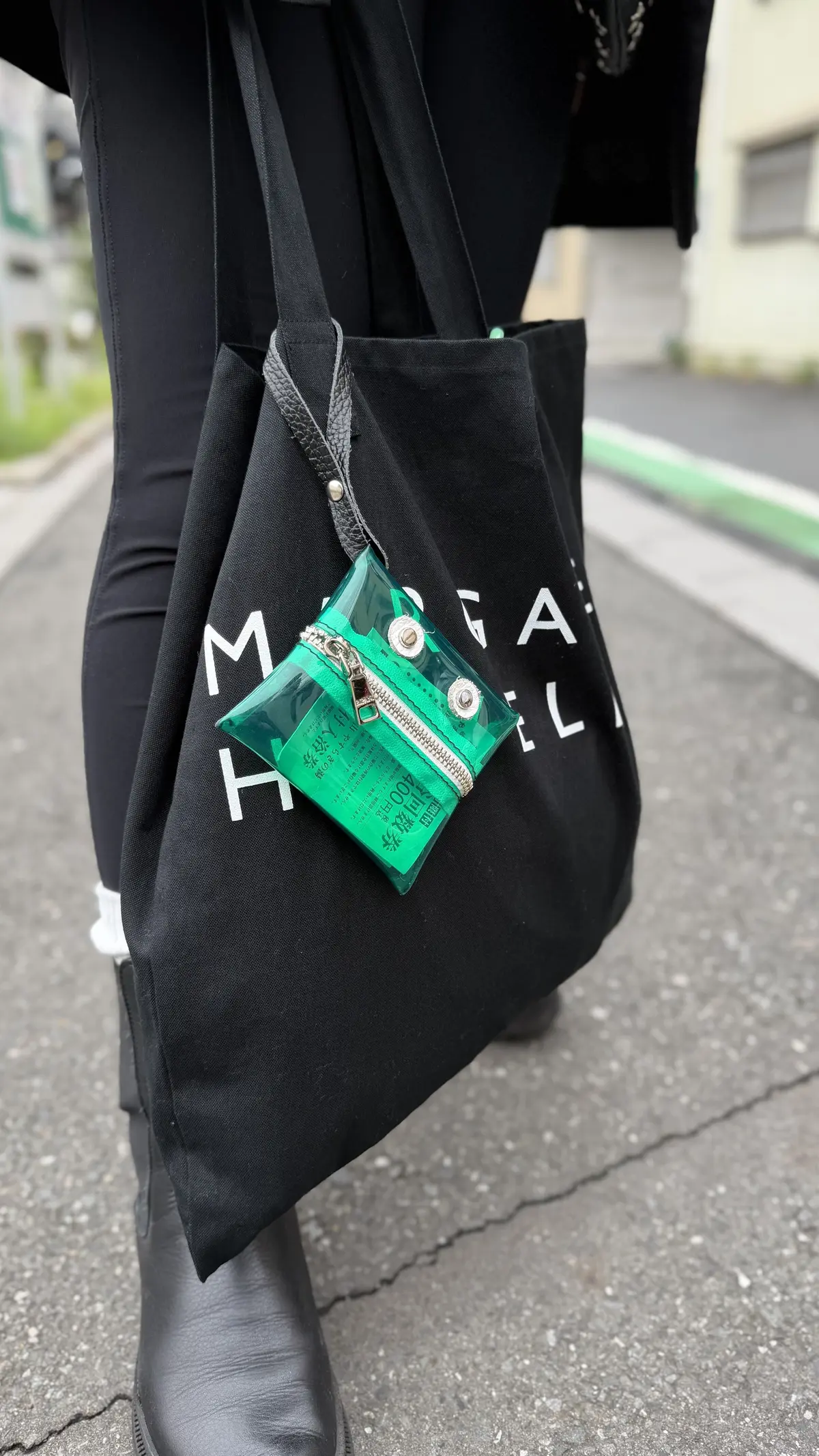 黒いトートバッグを肩にかけた人物。バッグには白い文字で「MEGA」とプリントされている。バッグの前面に、透明な緑色のポーチがぶら下がっており、中には日本の紙幣が見える。