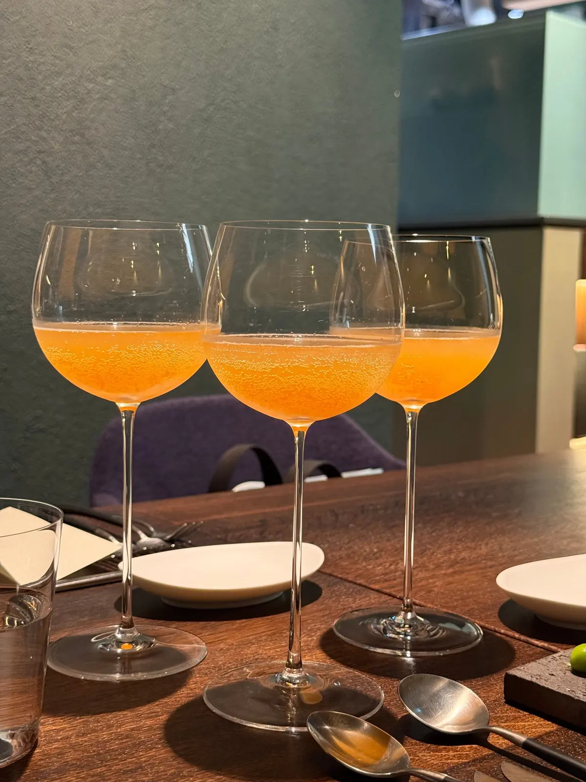 木製のテーブルに置かれた3つのワイングラス。グラスにはオレンジ色の炭酸飲料が入っており、泡が立っている。背景には緑色の壁と紫色の椅子が見える。