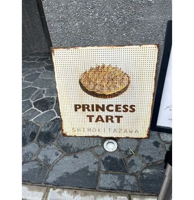 錆びた金属製の看板に、プリンセス タルトのイラストと文字が描かれている。背景はコンクリートの壁、足元は石畳。看板の下部に「SHIMOKITAZAWA」の文字。