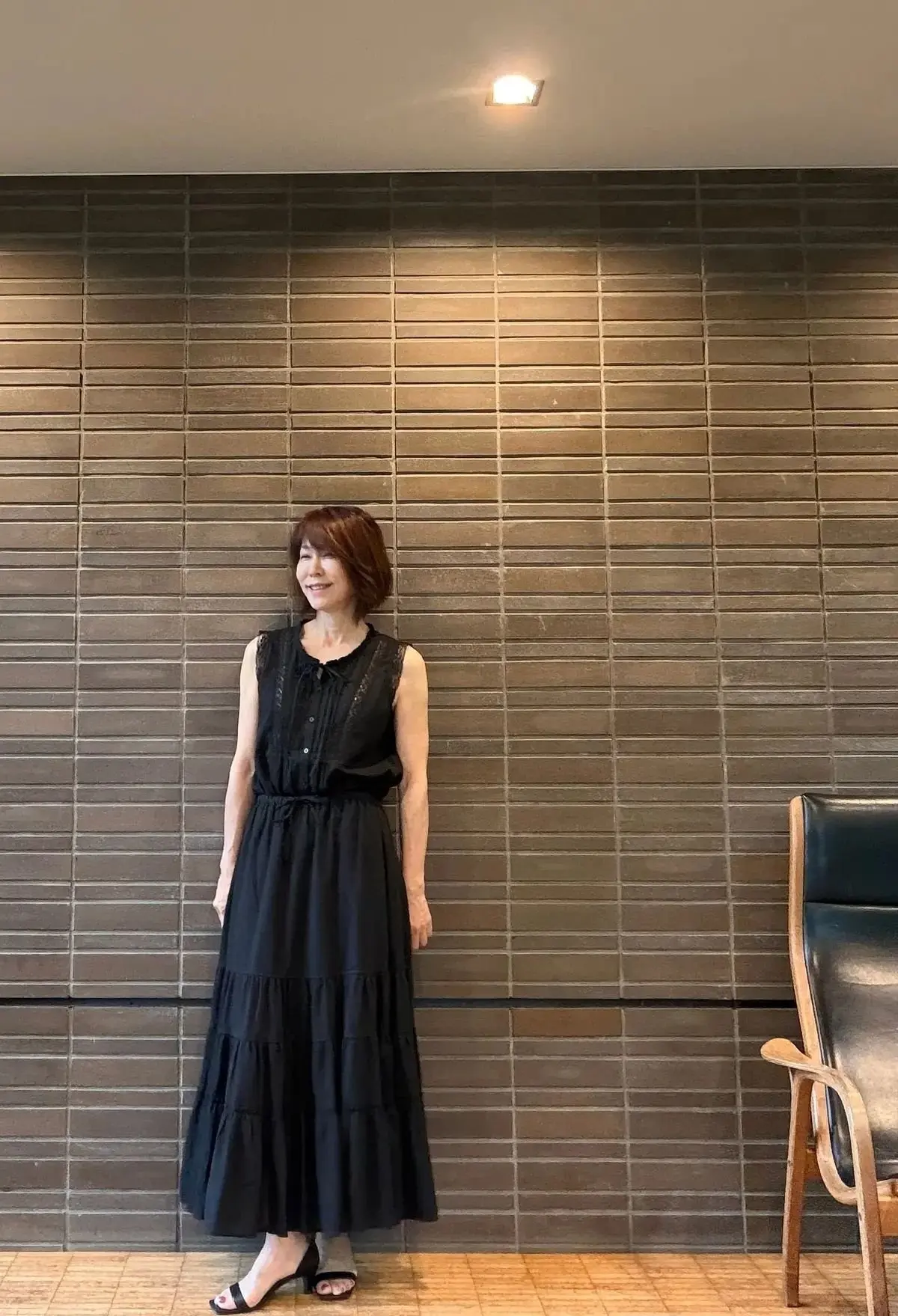 茶色のタイル壁に、黒のフレアスカートとトップスを着た女性が立っている。右側に木製の椅子がある。