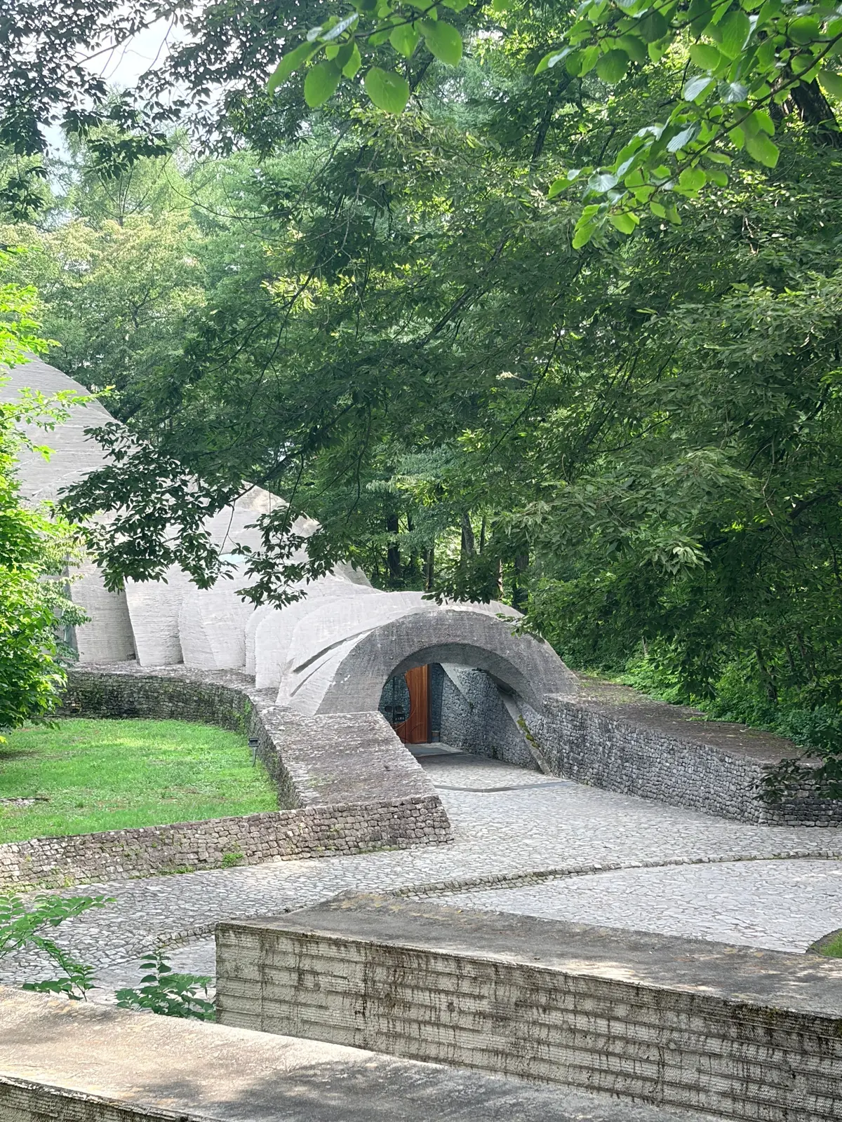 緑豊かな木々に囲まれた、コンクリートと石積みの建造物。アーチ型の入り口があり、奥には木製の扉が見える。手前に石畳の道と、緑の芝生が広がっている。
