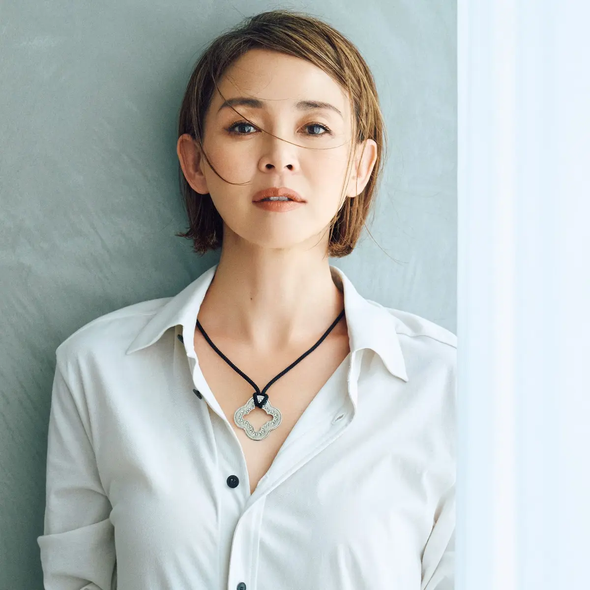 白いシャツを着た女性が、窓際に立ってこちらを見ている。短くカットした茶色の髪、控えめなメイク、銀色の装飾品ネックレスを身に着けている。背景は淡いグレーの壁。