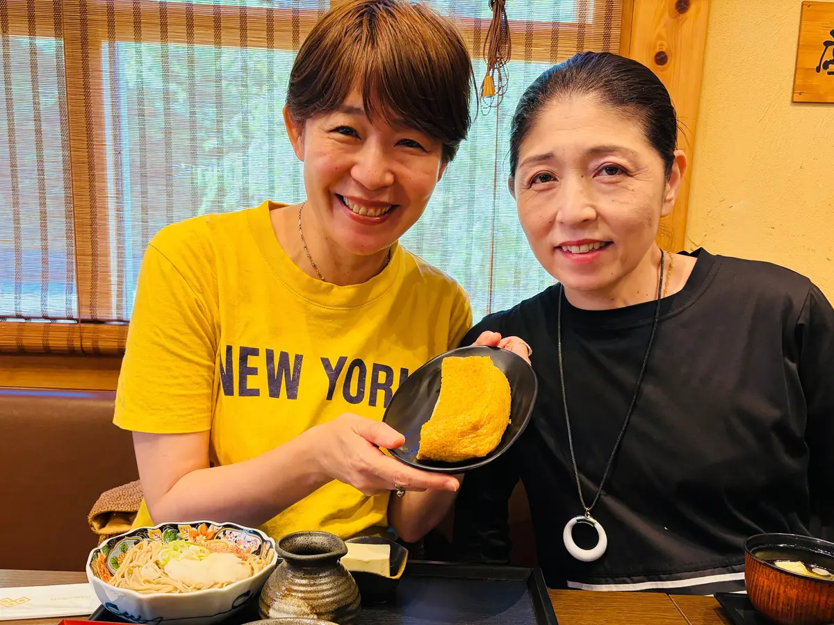黄色と黒の服を着た二人の女性が、木製のテーブルに置かれた料理を囲んで笑っています。中央には卵焼きがのった黒い皿があり、周囲にはサラダ、汁物、陶器の容器などが並んでいます。背景には窓と和風のインテリアが見えます。