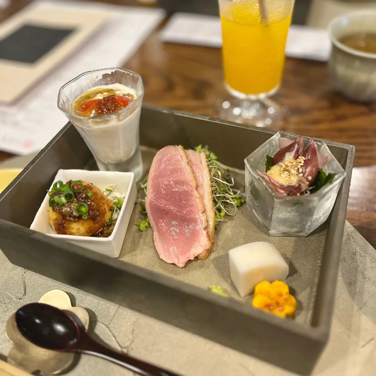 トレイに並べられた料理セットで、ピンク色の生肉、豆苗、小さな白い容器に入った揚げ物、ガラスの容器に入った前菜、オレンジジュースとコーヒーなどがあります。前菜盛り合わせ