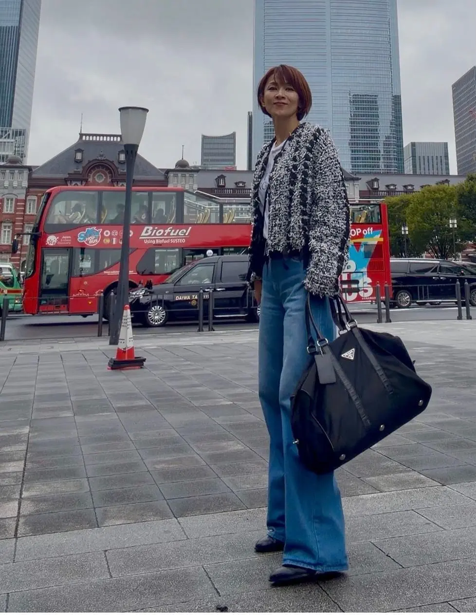 都会の街並みを背景に、黒いボストンバッグを持つ女性。青いフレアジーンズに黒いトップス、黒いパンプスを着用。茶色のショートヘアで、やや微笑んでいる。背景には赤い二階建てバスと高層ビルが見える。