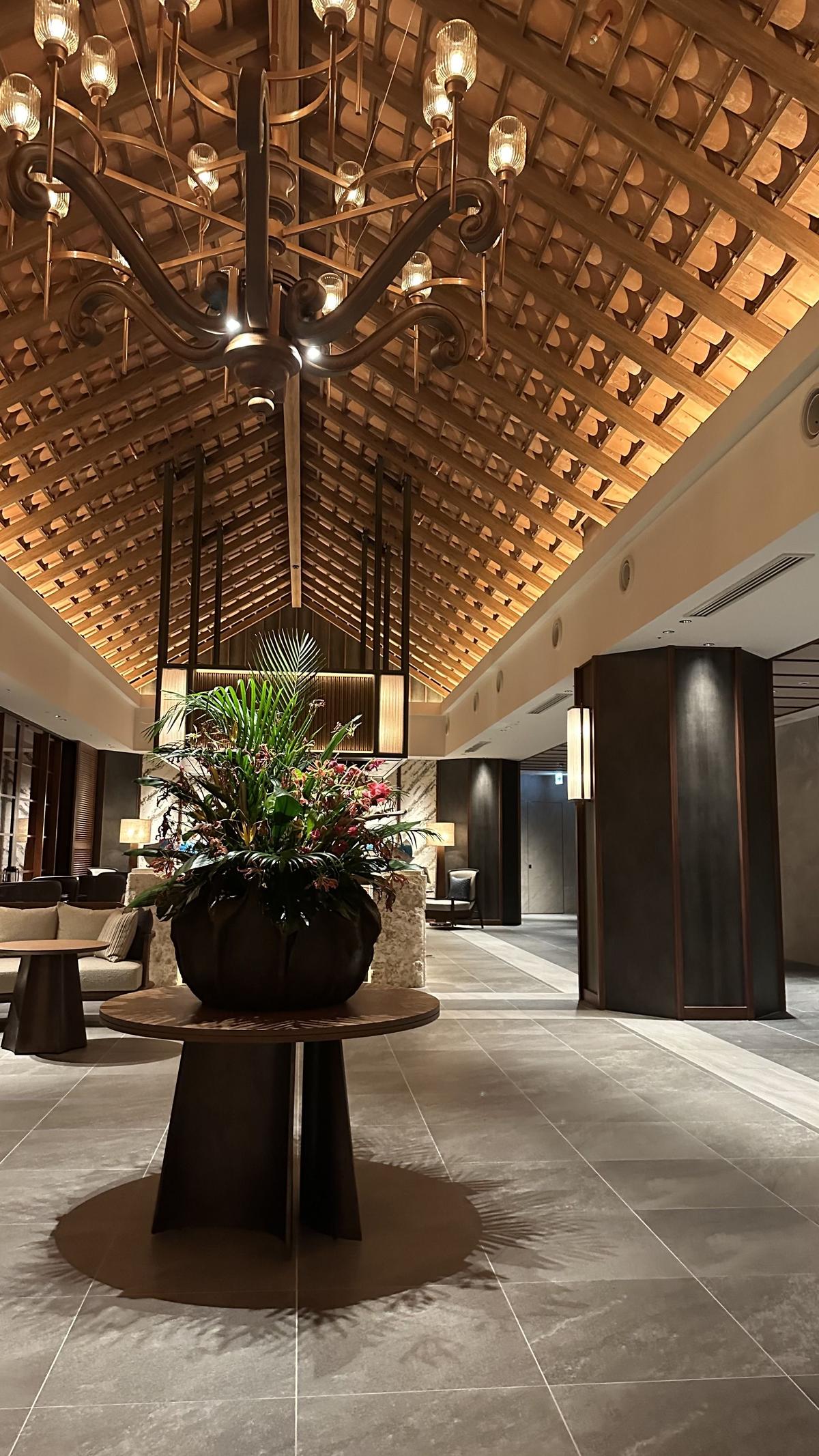 ホテルのロビー。中央に大きな植栽と丸いテーブルがあり、天井には木目調の梁と豪華なシャンデリアが設置されている。床はグレーのタイル張り。