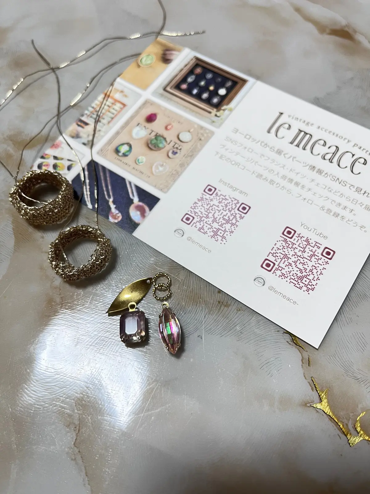 ビンテージアクセサリー「le meace」のジュエリーと販促資料。ゴールドのネックレス、リング、イヤリングが置かれ、QRコード付きの販促カードが中央に配置されている。背景は白いテーブル。