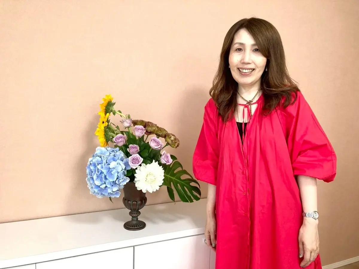 ピンクのワンピースを着た女性が、花の入った花瓶と共に明るい部屋に立っています。