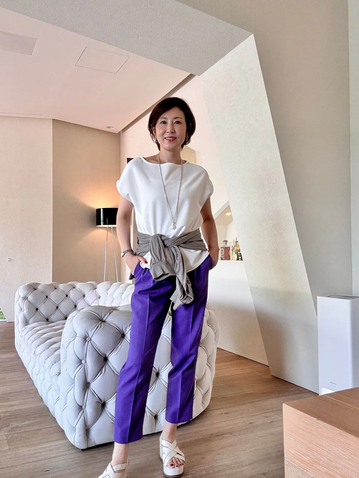 白のトップスに紫のパンツを着用した女性が、モダンな内装の部屋でポーズをとっている。右手に手を置き、白いソファの前に立っている。