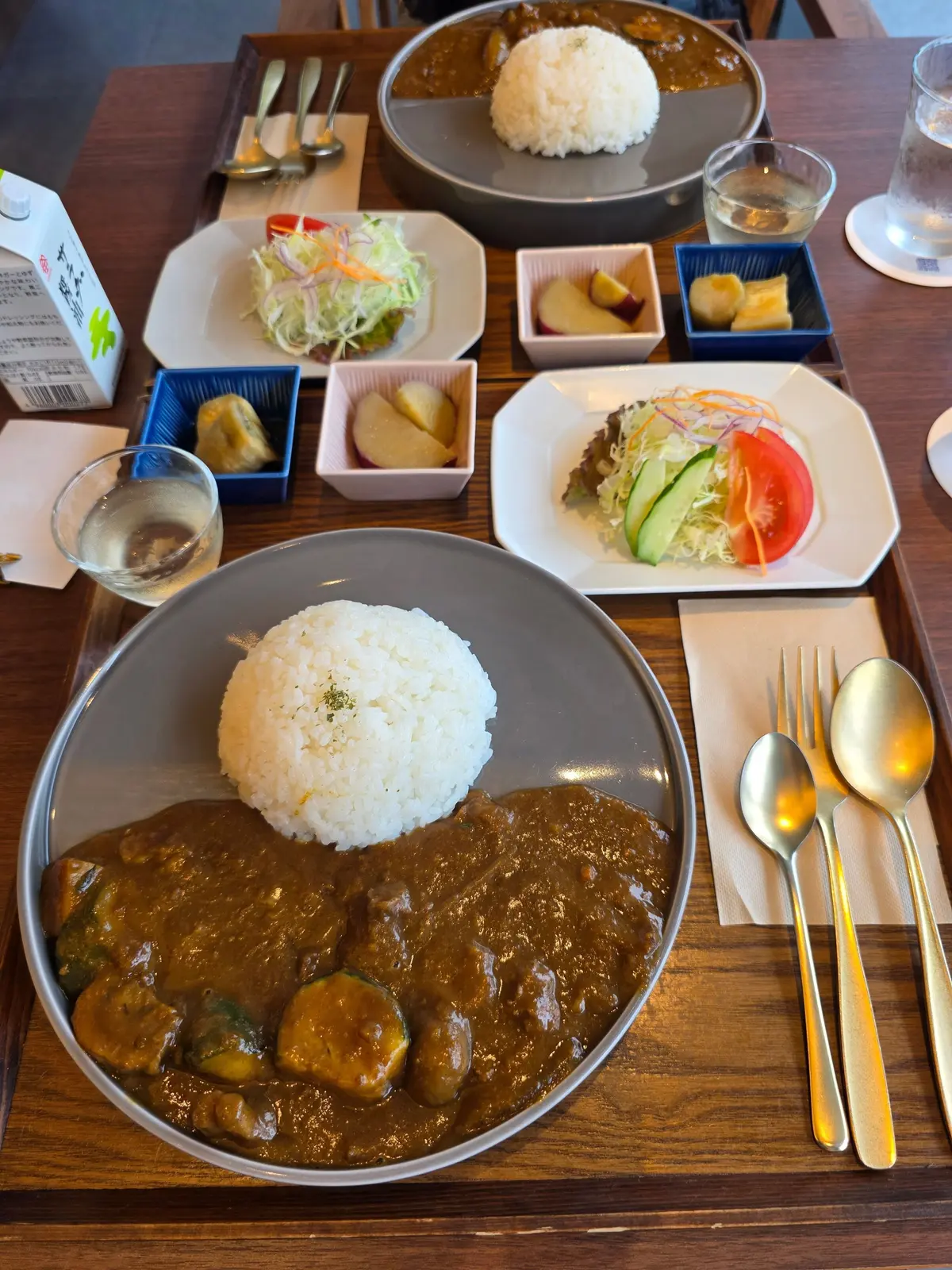 木製のトレイに盛り付けられた日本の定食。中央に白米とカレー、右にはサラダや漬物などの小鉢が並び、飲み物とカトラリーも添えられている。