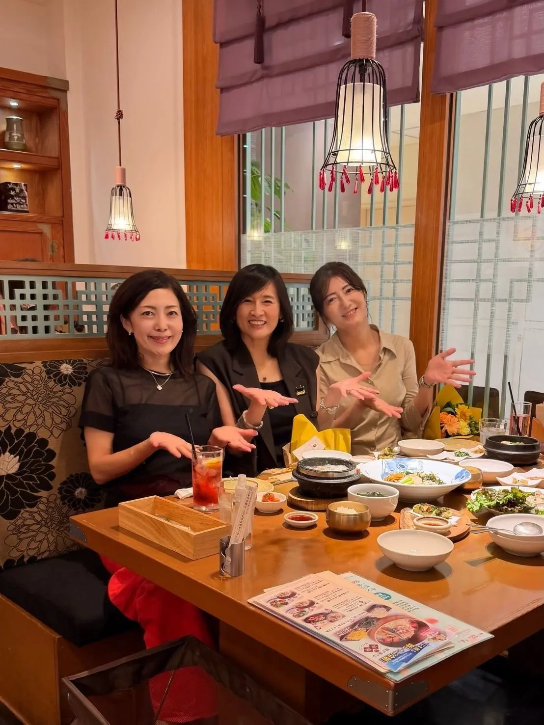 レストランで食事を楽しむ3人の女性。笑顔で手を広げ、テーブルには様々な料理と飲み物が並んでいる。背景には和風の照明と内装が見える。
