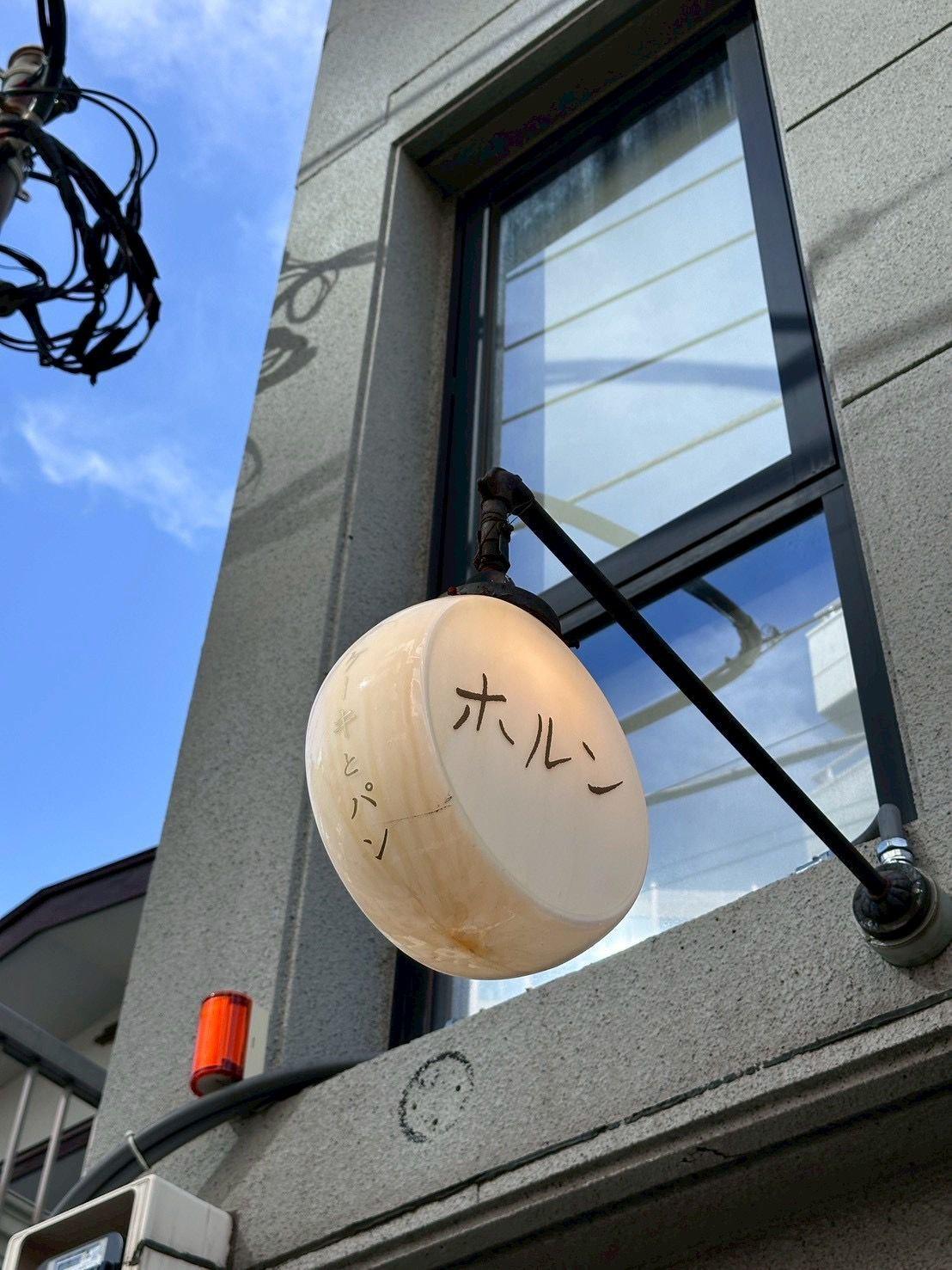 コンクリートの壁に設置された球体の照明器具。表面には黒い文字で「ホールン」と書かれている。窓の反射が映り、背景には青空が広がっている。