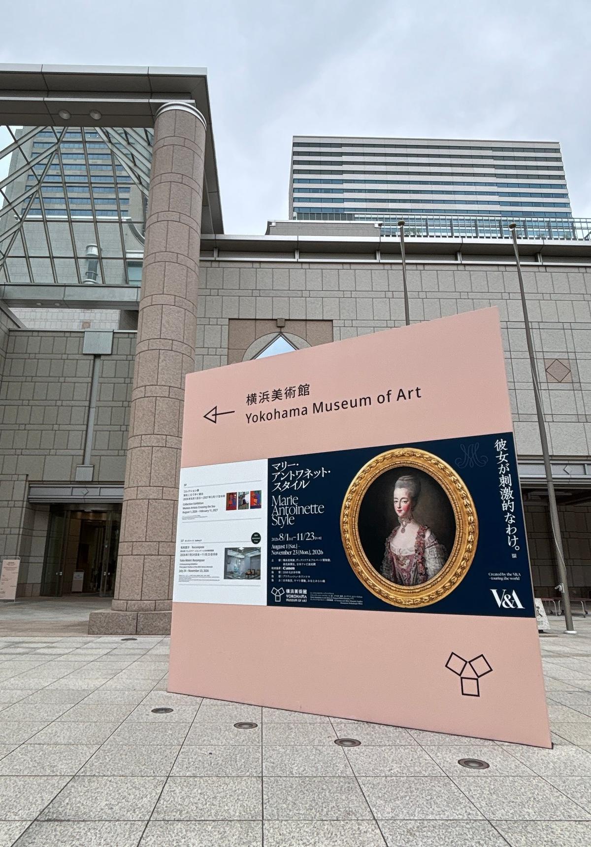 横浜美術館の外観と、展示案内の看板。近代的なコンクリートの建物で、右側に「Yokohama Museum of Art」の文字。看板には「Marie Antoinette Style」の展示案内の写真とテキスト。右奥には縦長の看板が立っている。